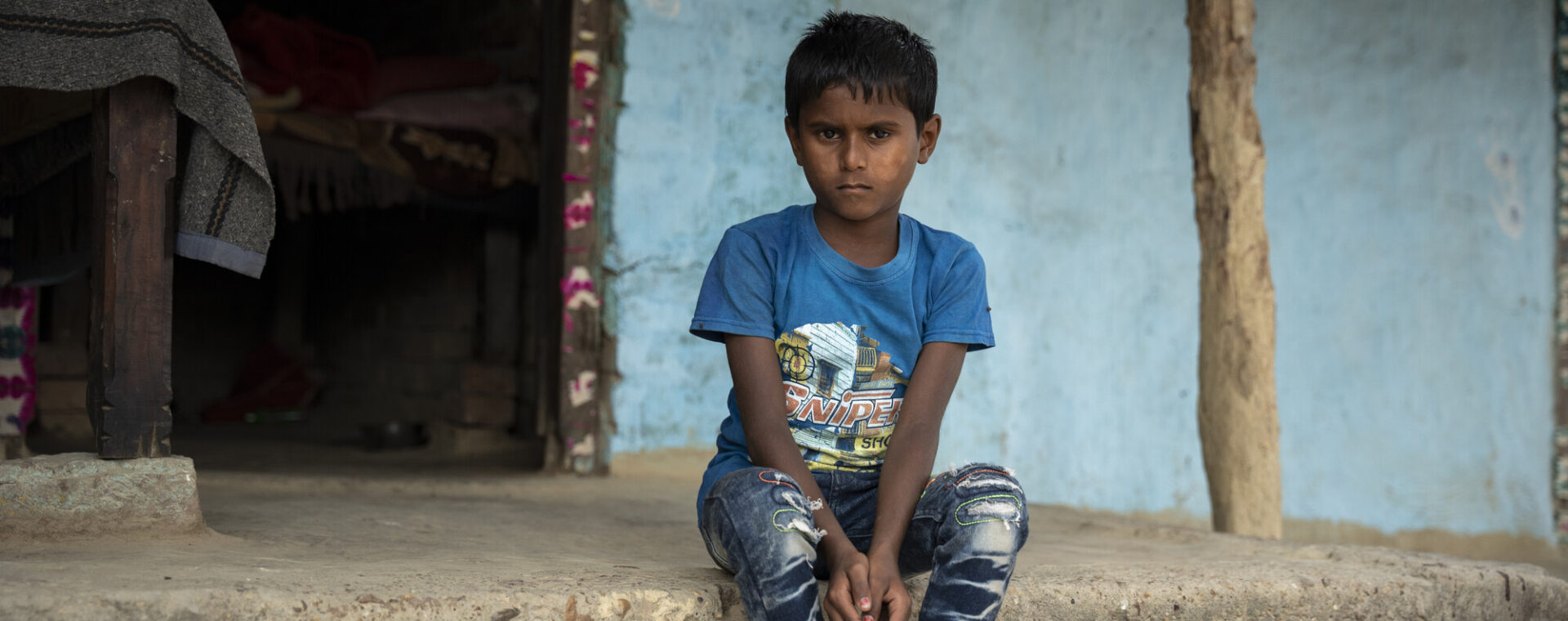 A little boy wearing a t-shirt and jeans sits on the porch of a blue painted house.