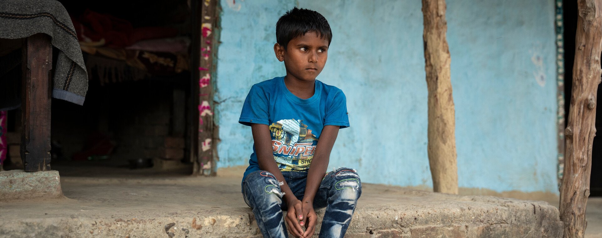 A little boy wearing a t-shirt and jeans sits on the porch of a blue painted house.