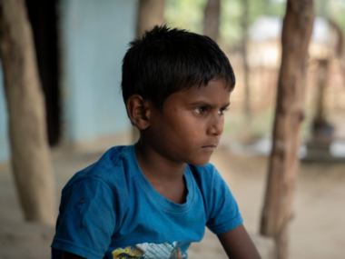A little boy wearing a t-shirt sits on the porch of a blue painted house and looks off to the right.