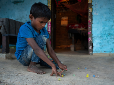 A little boy wearing a t-shirt crouches on the porch of a blue painted house, playing with marbles.