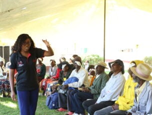 A group of people wearing eye patches following cataract surgery are sitting down. Stood in front of them is Felistas from CBM partner HelpAge Zimbabwe.
