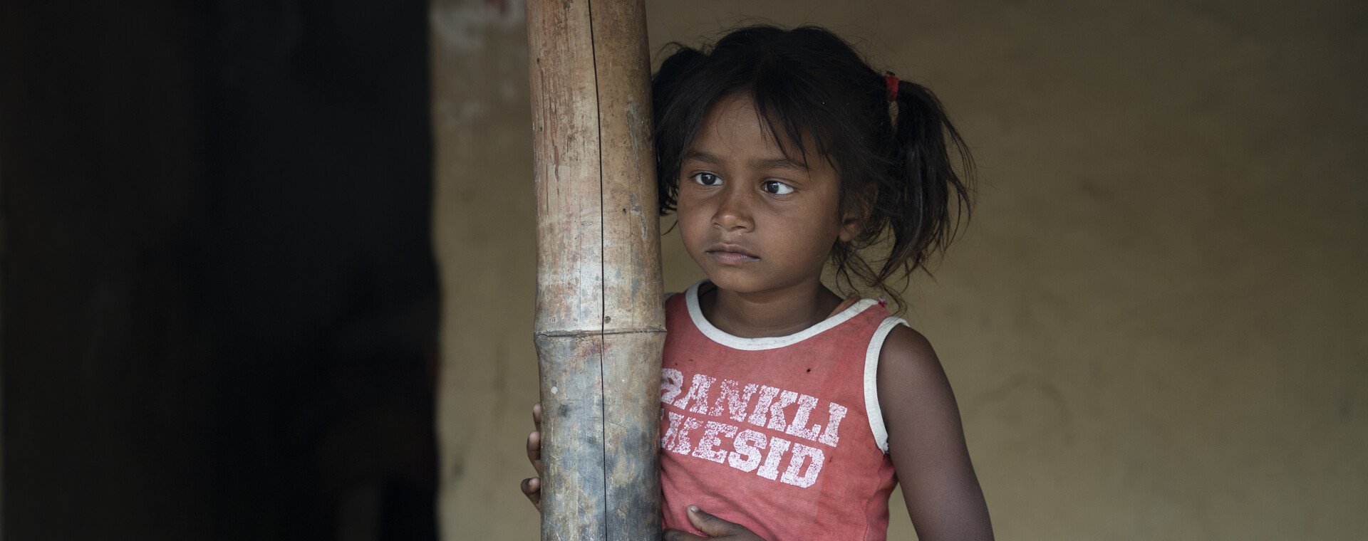 A little girl with dark hair and a red and white t-shirt stands outside her house and leans against a wooden post.