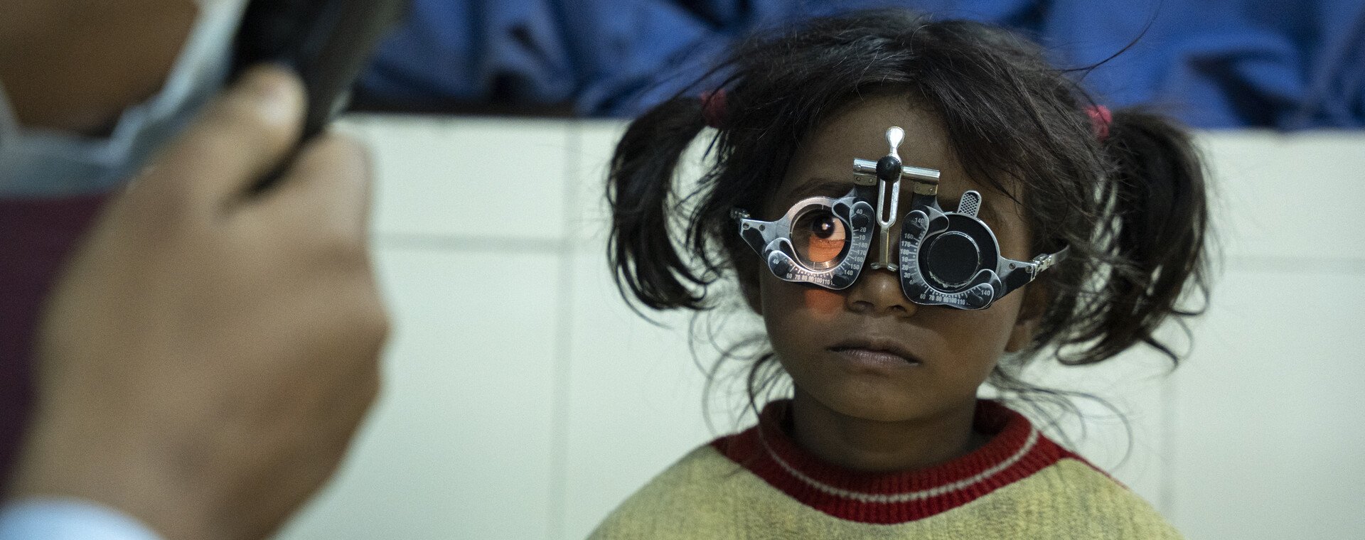 A little girl with dark hair, wearing a cream and red jumper is wearing a pair of eye test glasses. She looks at a doctor, whose hand can be seen in the left corner, as he assesses her eyes.