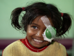 A little girl with dark hair and a cream and red jumper smiles into the camera. She has a green patch on her left eye.