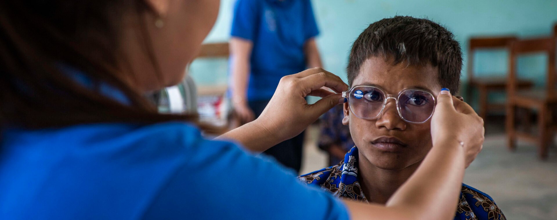 A boy in a classroom, is having glasses put on his face by a refractionist.