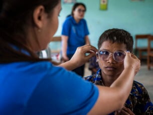 A boy in a classroom, is having glasses put on his face by a refractionist.
