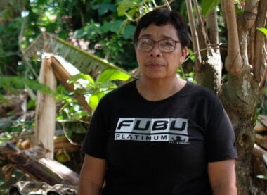 A lady wearing a black t-shirt stands outside her home. Behind her are trees and a house.
