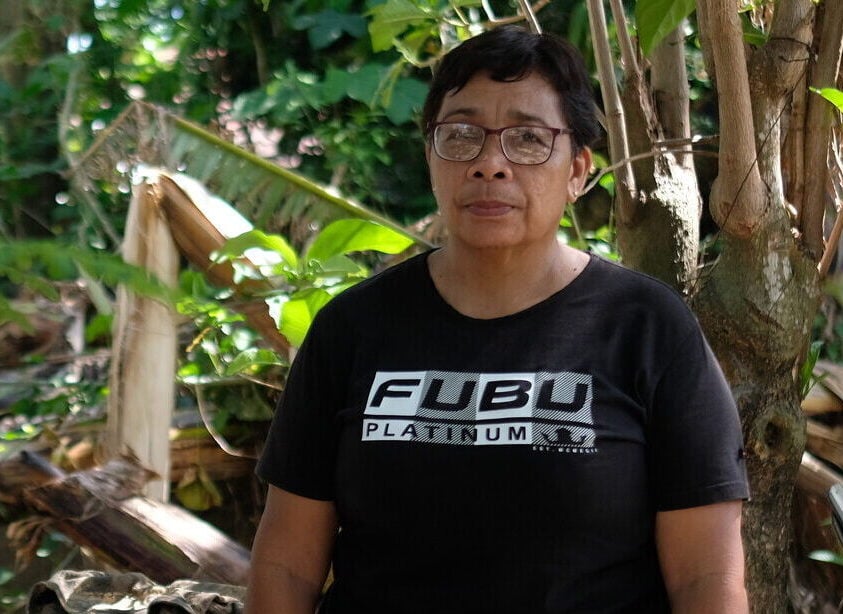 A lady wearing a black t-shirt stands outside her home. Behind her are trees and a house.