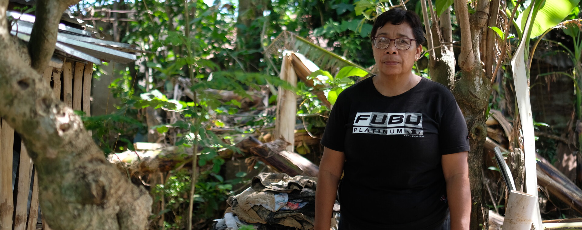 A lady wearing a black t-shirt stands outside her home. Behind her are trees and a house.