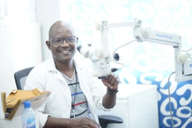 Henry, wearing a white coat sits in his chair behind some optical equipment.