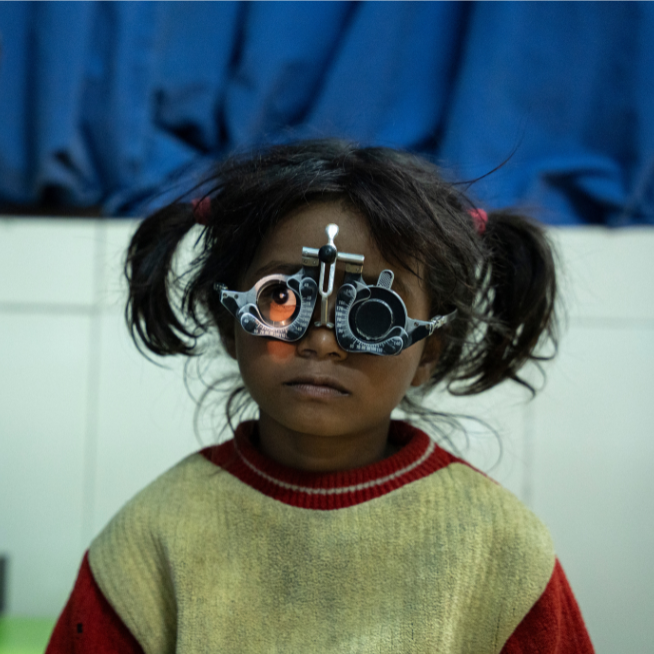 A little girl with dark hair and a cream and red jumper wears eye test glasses as a light is shone in her right eye.