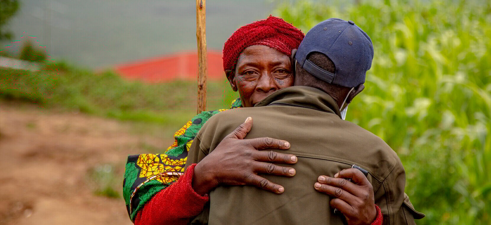 A lady wearing a colourful top and red headscarf, and a man wearing a brown jacket and blue hat embrace each other in a hug.