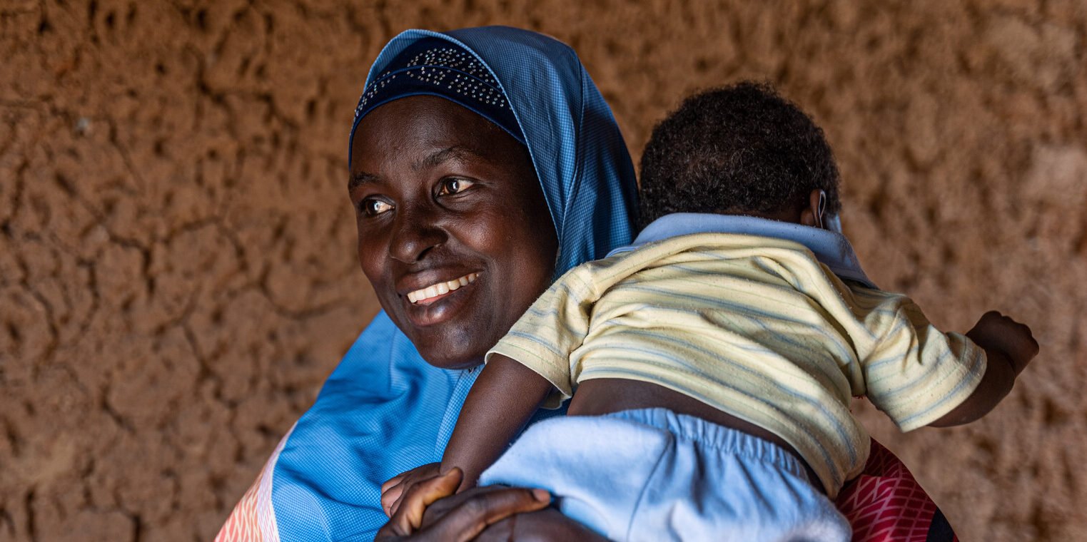A woman wearing a blue head scarf smiles and looks to the left, she holder her baby, who is wearing blue shorts, and a yellow t-shirt.