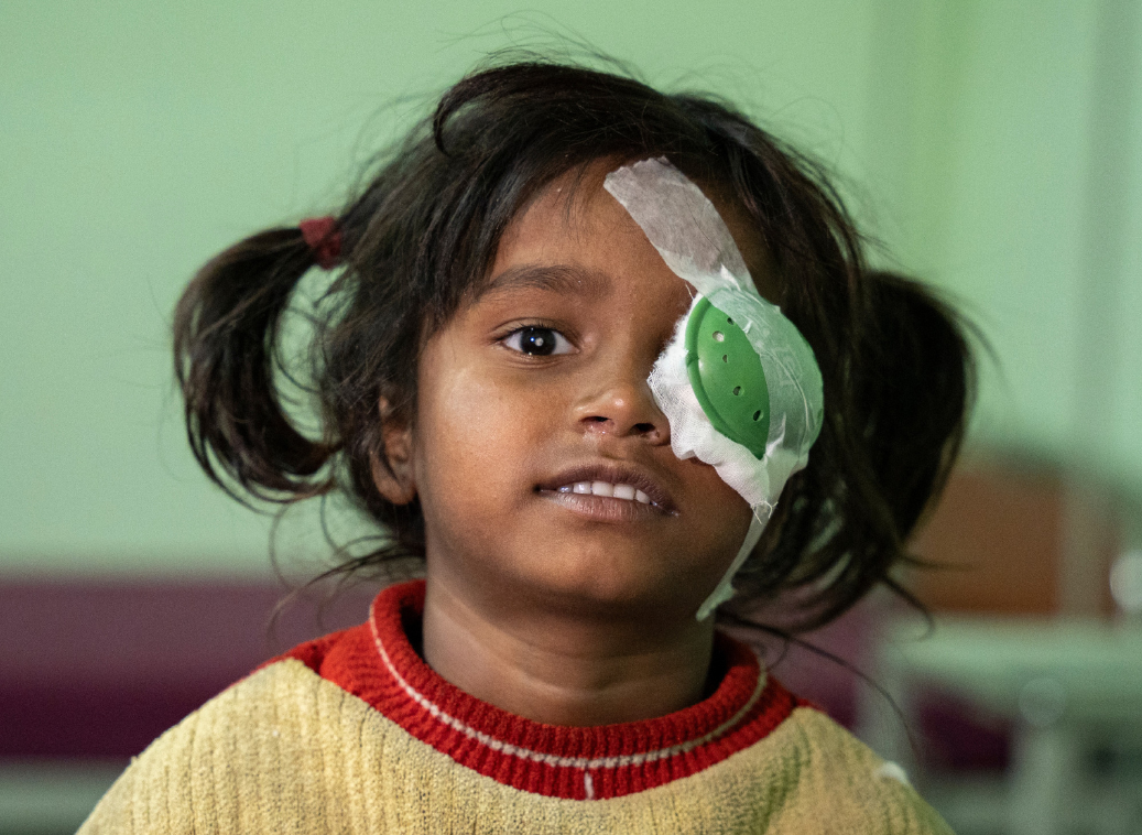 A little girl with dark hair and a cream and red jumper smiles into the camera. She has a green patch on her left eye.