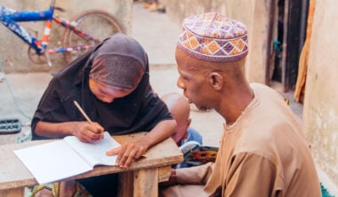 A girl wearing a hijab leans on a bench and writes in an exercise book whilst a man in a purple and orange hat sits beside her.