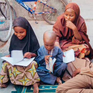 Three children sit on the ground looking at books. A little girl wearing a brown sari, another girl wearing a navy sari and patterned skirt, and a little boy wears a blue top and trousers.