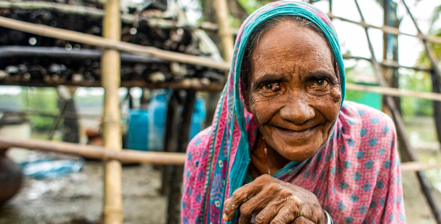 Khadija stands, resting on a walking stick and smiles at the camera. Behind her is a wooden structure.