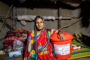 Rajida, wearing red, sits down, resting her arm on a red container.