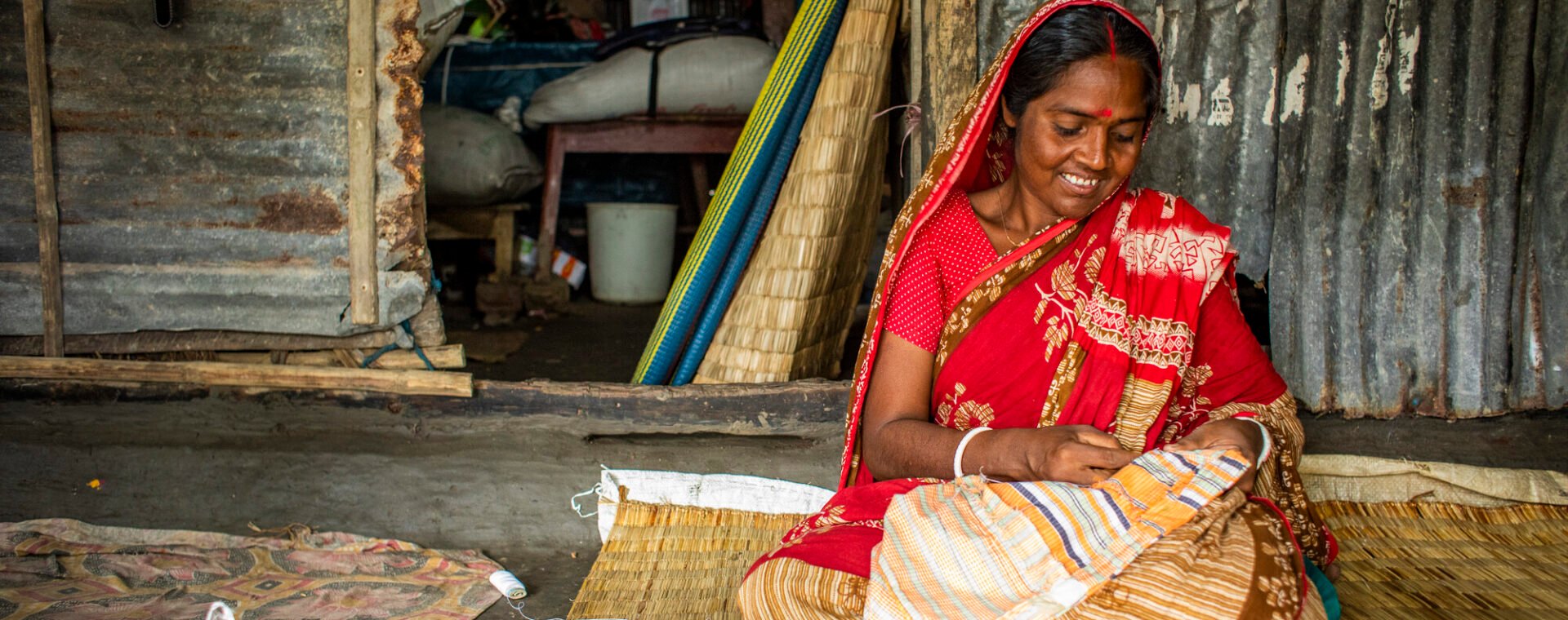 Topati, wearing red, sits on the floor and sews.