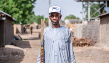 A man in a pale blue shirt and a white baseball cap, with a serious expression, holding a long ruler for measuring children's growth.