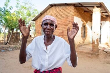 Dorothy standing in front of her house, wearing a white shirt and waving her hands around in joy.
