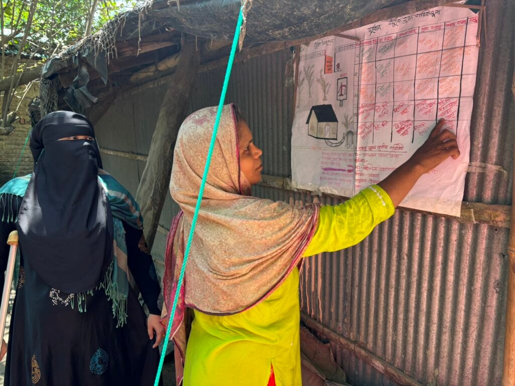 A woman, points at a chart taped to a wall while another woman stands behind her watching on.