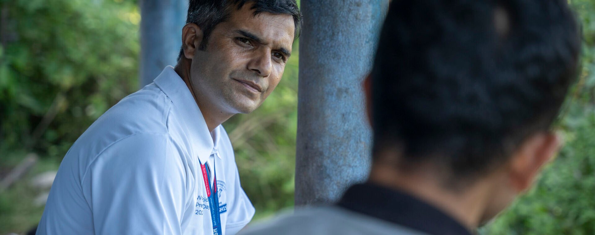 A man sits down, listening to another man who has his back to the camera.