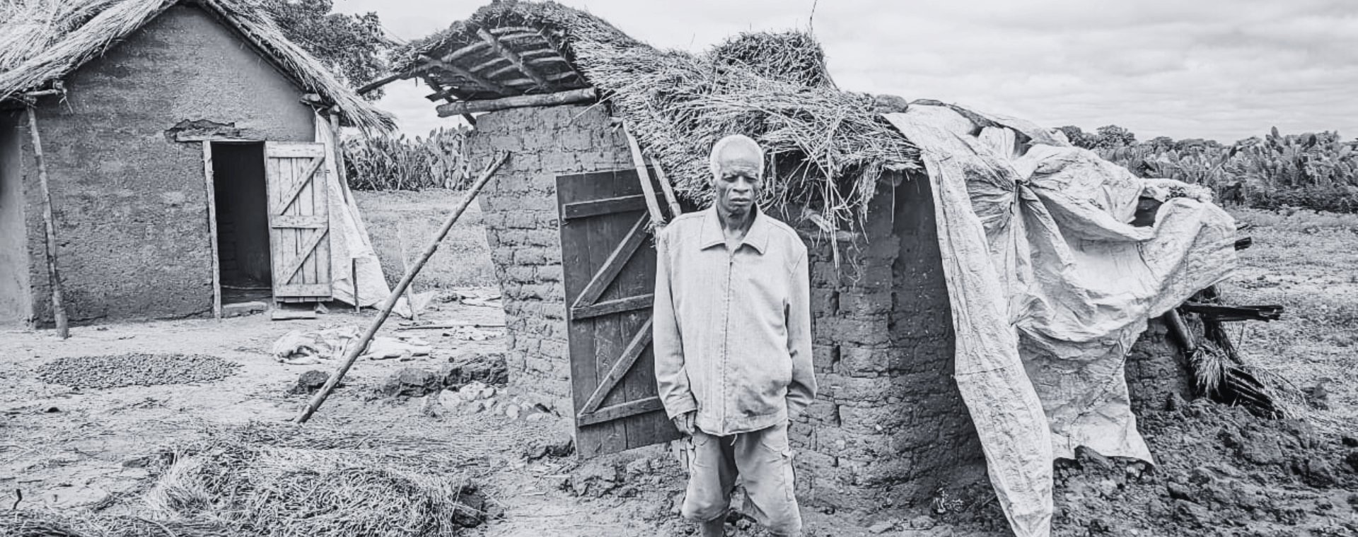 Black and white image of a man standing in front of a damaged brick hut.