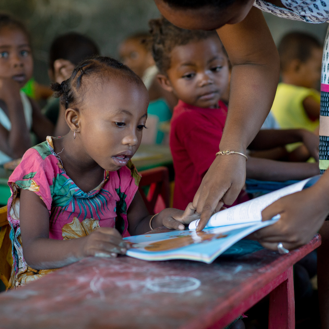 A little girl is sitting at a school desk. She is wearing a pink t-shirt with yellow and blue leaves printed. A teacher holds a book to her and points on the page.