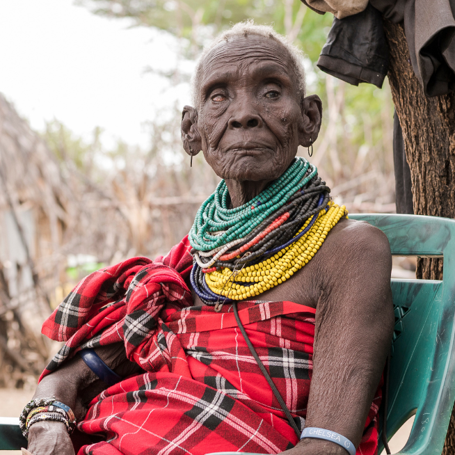 A lady is wearing a red scarf dress and red, blue and yellow beaded necklaces around her neck. She is sitting on a chair and looks into the camera.