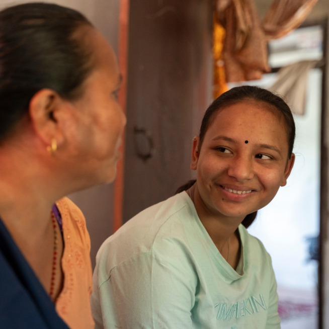 A teenage girl wearing a light green t-shirt smiles to her mother on the left hand side of the photo. Her mother wears an orange and black dress.
