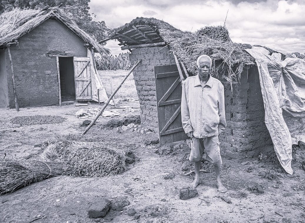 Black and white image of a man standing in front of a damaged brick hut.