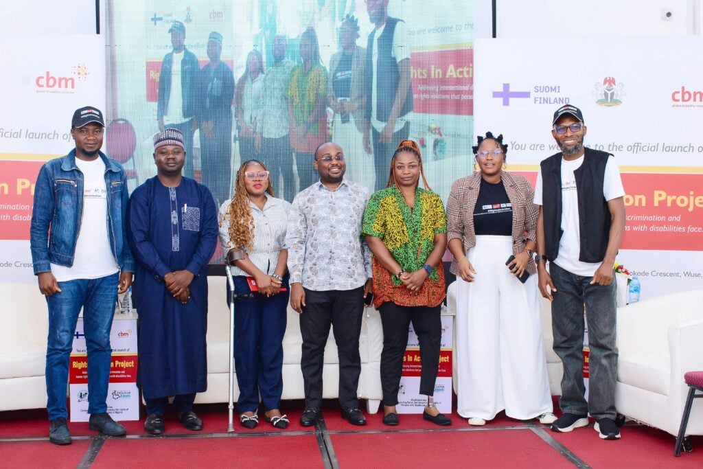 Several people are stood on a stage at the Rights in Action project launch in Nigeria. There is a screen and banners behind them.