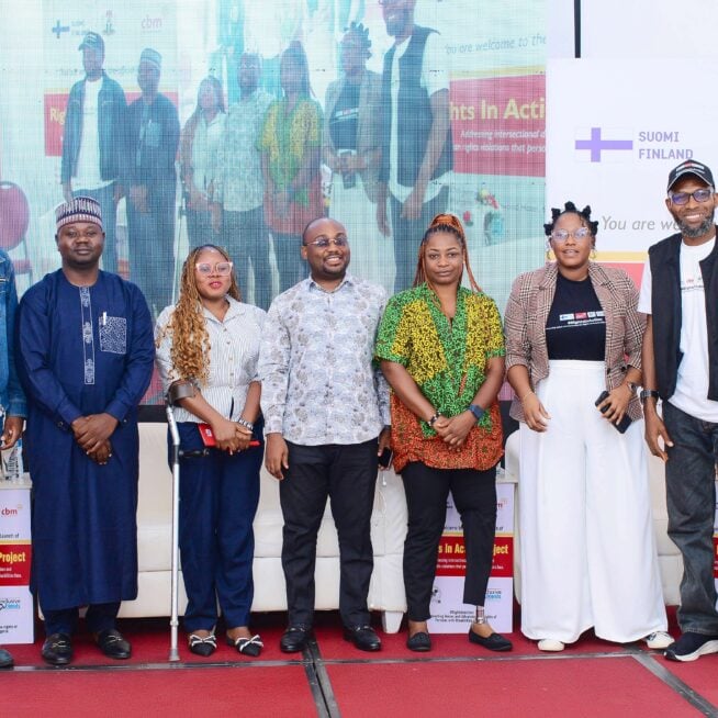 Several people are stood on a stage at the Rights in Action project launch in Nigeria. There is a screen and banners behind them.