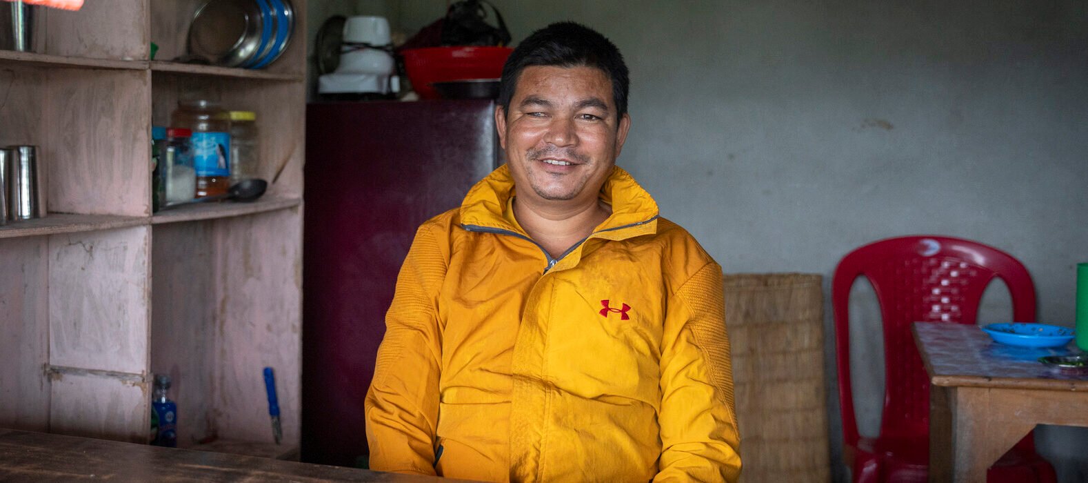 A man wearing a yellow coat sits on a chair at a desk inside a shop. He looks into the camera and smiles, his right eye visible with cloudiness.