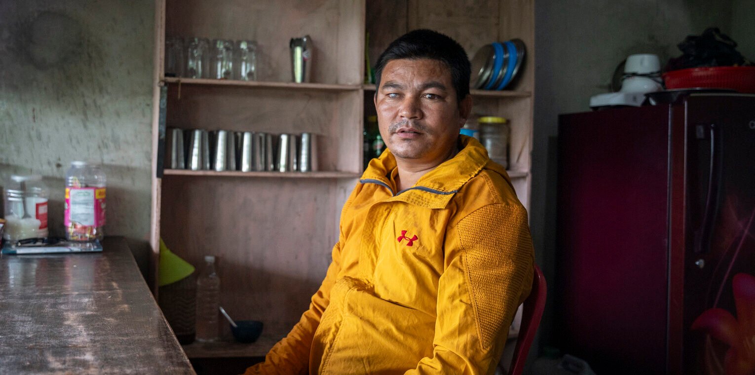 A man wearing a yellow coat sits on a chair inside a shop. He looks into the camera, his right eye visible with cloudiness.