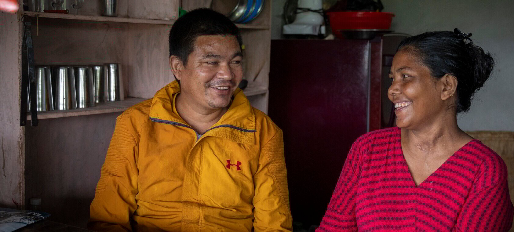 A man wearing a yellow coat sits on a chair inside a shop. He smiles at a lady next to him, his right eye visible with cloudiness. A lady in a red and black top sits on a chair next to the man, and smiles to him.
