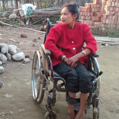 A lady wearing a red knitted cardigan and black trousers sits in a wheelchair. She is outside in amongst rocks and bricks. She looks to her right.