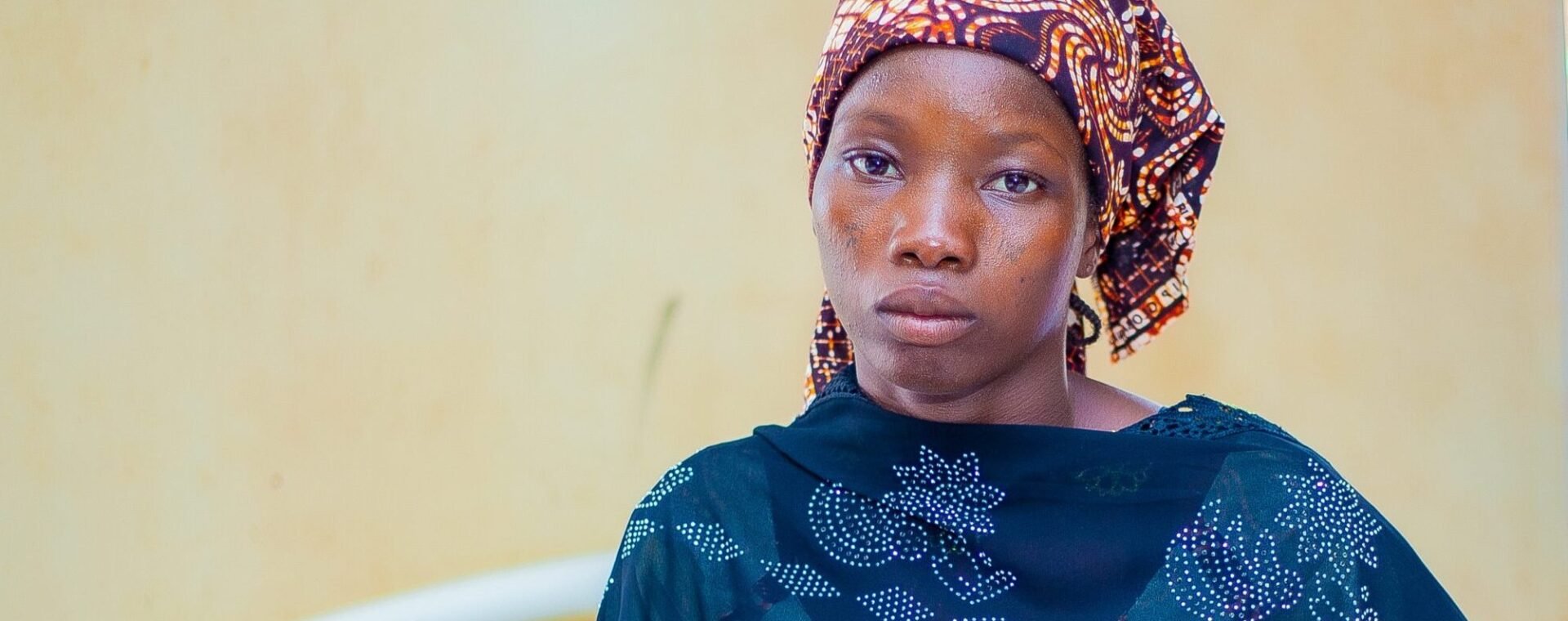 A Nigerian woman wearing a brown patterned headscarf and a dark blue top. She has a neutral expression on her face.