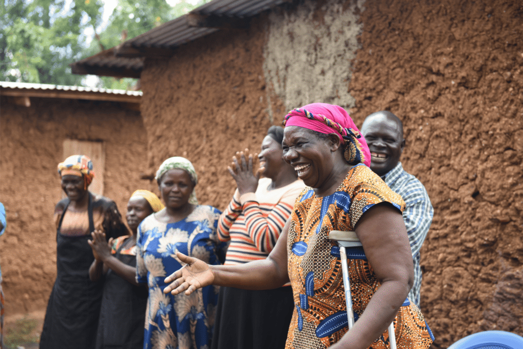 A group of people stood outside smiling and laughing. The woman who is standing closest to the camera has her hand outstretched and the other holding a crutch.