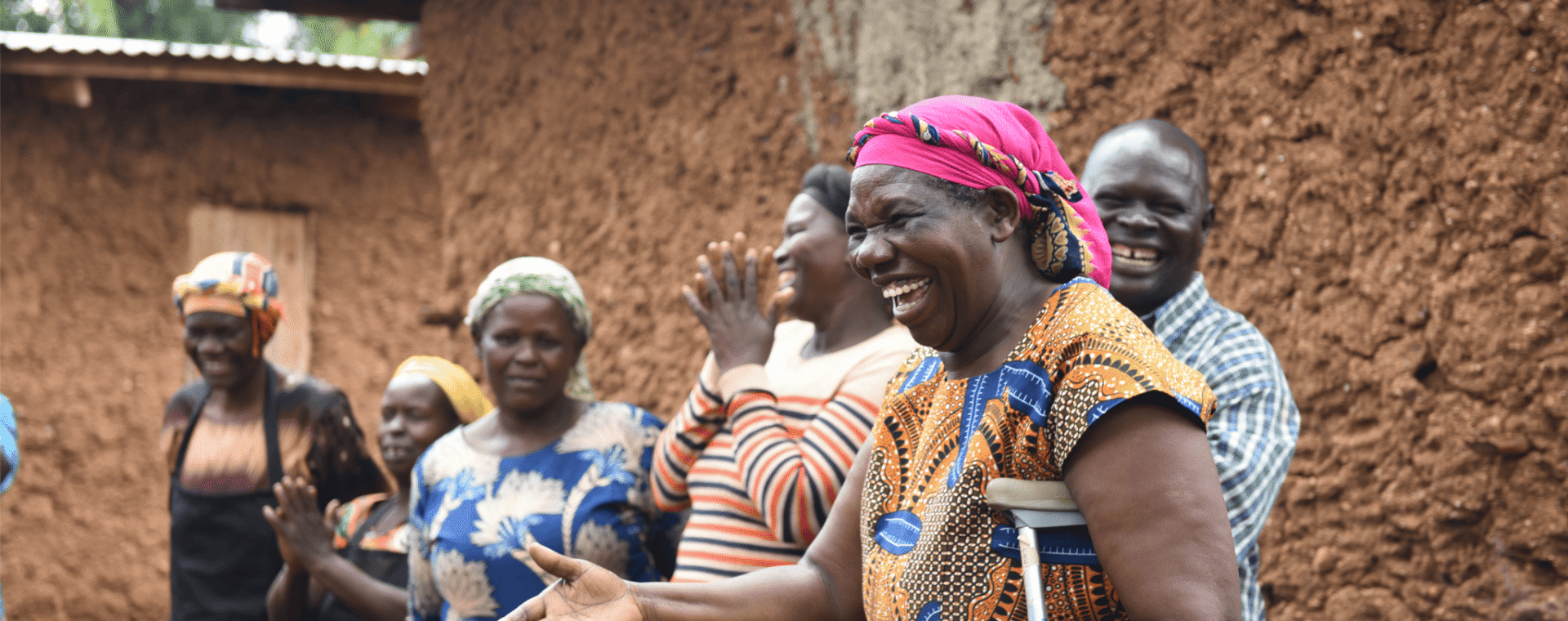 A group of people stood outside smiling and laughing. The woman who is standing closest to the camera has her hand outstretched and the other holding a crutch.