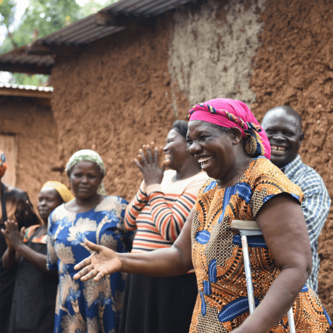 A group of people stood outside smiling and laughing. The woman who is standing closest to the camera has her hand outstretched and the other holding a crutch.