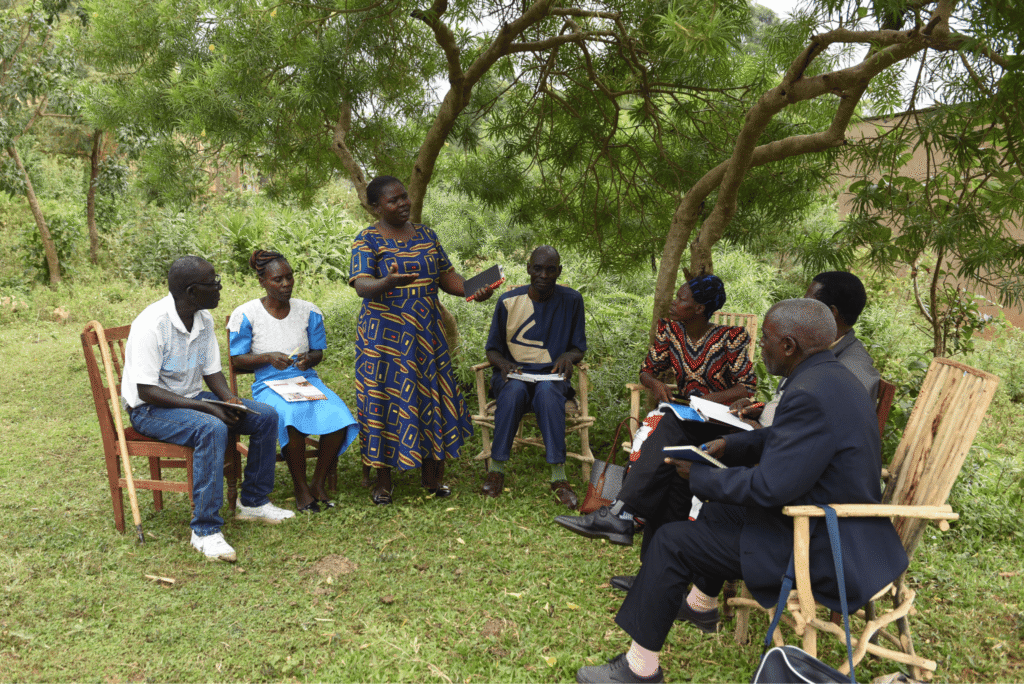 The members of a Local Transformation Unit are sat down outside, listening to one member who is stood up.