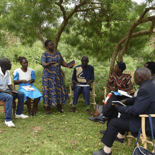 The members of a Local Transformation Unit are sat down outside, listening to one member who is stood up.