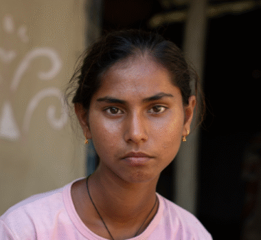 A girl, with dark hair pulled back, wearing a pink t-shirt, looks into the camera. She sits in the courtyard of her home.