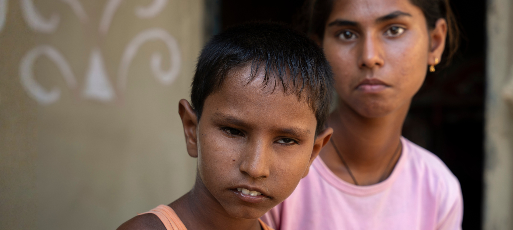 A little boy with dark, short hair, wearing an orange vest sits to the left of a girl, with dark hair pulled back, wearing a pink t-shirt. They sit in the courtyard of their home.