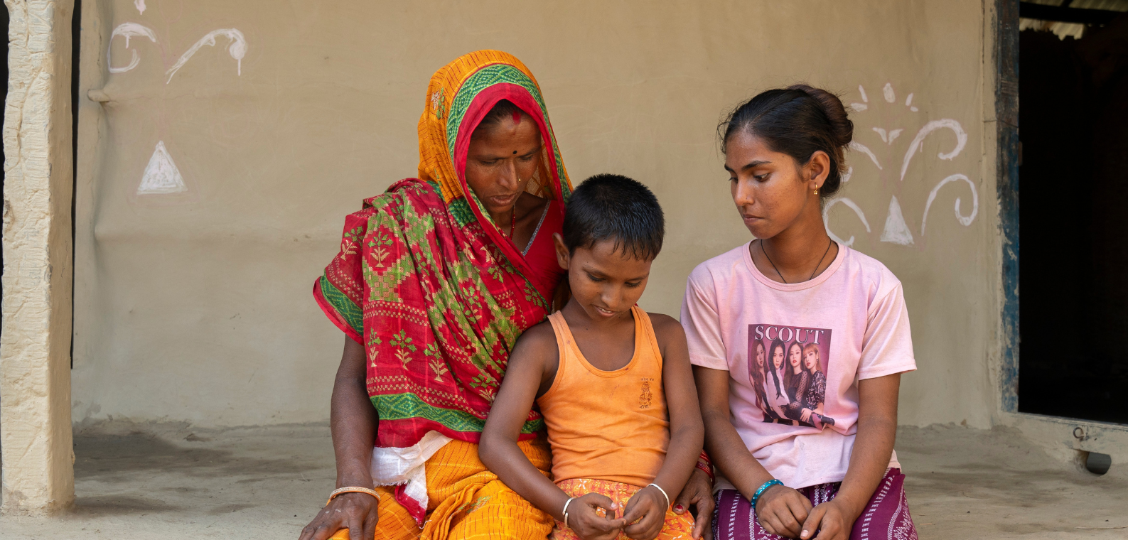 A little boy with dark, short hair, wearing an orange vest sits between his mother and sister. Pictured to his left is his mother, wearing an orange, green and red sari and dress. To his right, his sister, with dark hair pulled back, wearing a pink t-shirt and pink patterned trousers. They sit in the courtyard of their home.
