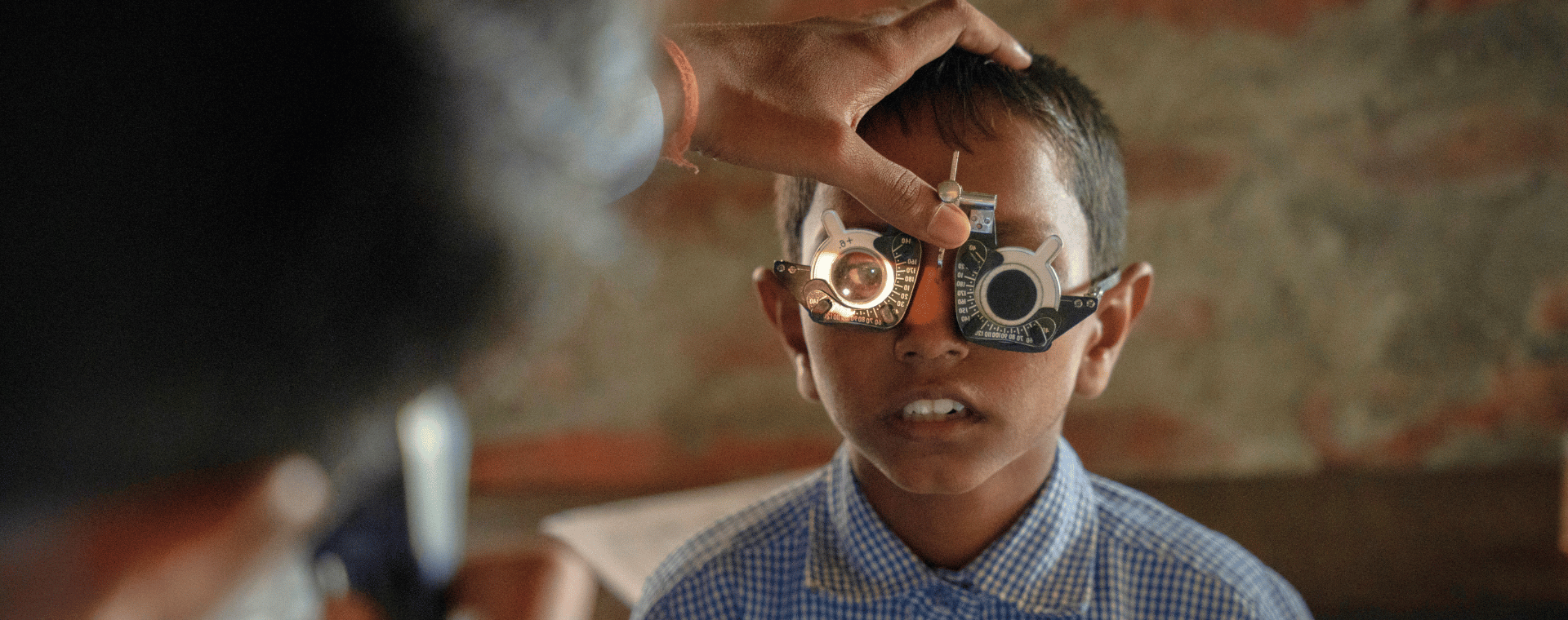 A little boy wearing a blue shirt wears a pair of eye examination glasses. A man with dark hair can be seen shining a light into the boys right eye while he holds the boys head still.