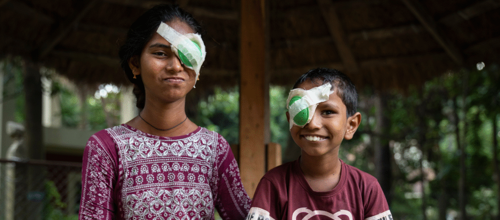 A girl wearing a pink and white patterned dress is sat outside under a hut smiling into the camera. She has dark hair pulled back, and has a green eye patch on her left eye. To her left is her brother, wearing a brown and white t-shirt, with short, dark hair, he has a green eye patch on his right eye. This image is being used as part of our Big Give Christmas Challenge.