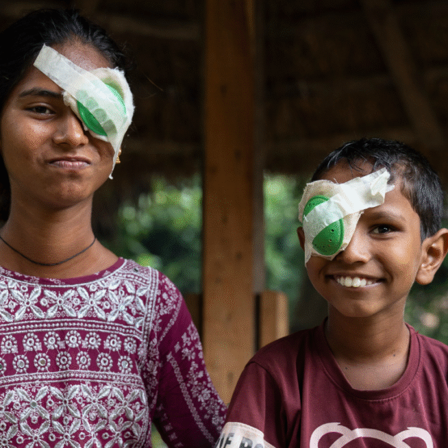 A girl wearing a pink and white patterned dress is sat outside under a hut smiling into the camera. She has dark hair pulled back, and has a green eye patch on her left eye. To her left is her brother, wearing a brown and white t-shirt, with short, dark hair, he has a green eye patch on his right eye. This image is being used as part of our Big Give Christmas Challenge.